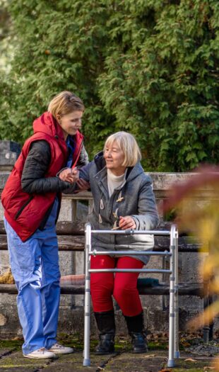 Senior woman with walking frame and caregiver outdoors sitting on bench in park, helping to stand up.